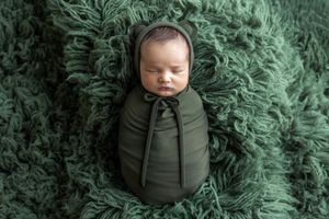 Newborn baby swaddled in olive green wrap and bonnet, peacefully sleeping on a lush green flurry backdrop.