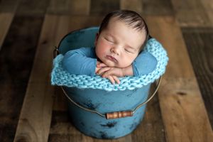 Sleeping newborn baby nestled in a rustic blue bucket with chunky knit blanket on wooden floor.