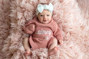 Baby girl in pink knit romper with name Lainey, white bow headband, lying on fluffy pink rug.