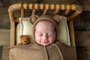 Smiling newborn baby in brown bear bonnet sleeping in miniature wooden bed with teddy bear prop.