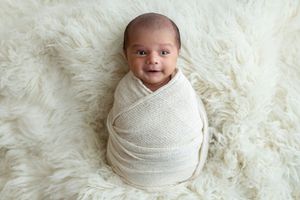 Smiling newborn baby swaddled in cream knit wrap lying on fluffy white fur blanket.