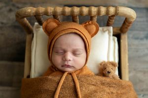 Sleeping newborn baby wearing a brown bear bonnet wrapped in a matching knit blanket in a wooden crib.