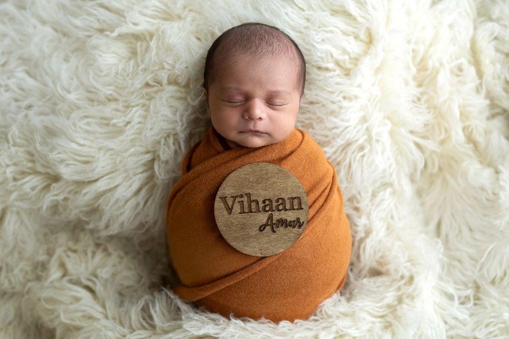 Newborn baby Vihaan Amor swaddled in burnt orange wrap on fluffy white blanket with wooden name disc.