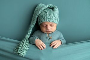 Sleeping newborn baby wearing a teal knitted stocking cap and matching romper, posed against a teal background.