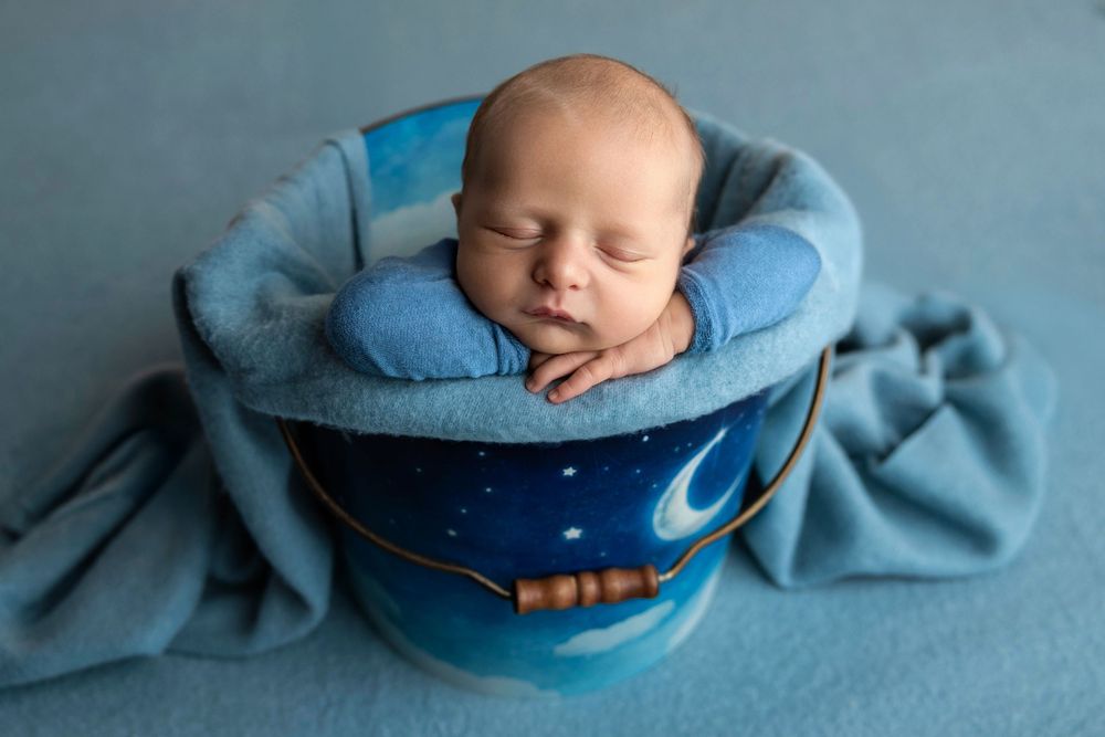 Newborn baby sleeping peacefully in a blue decorative bucket with a soft blue blanket.