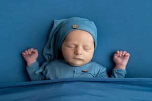 Newborn baby sleeping peacefully in blue outfit and hat on blue background with tiny fists raised.
