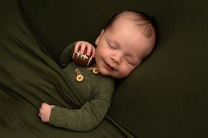 Sleeping newborn baby in green romper holding a tiny football on a green backdrop.