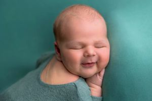 Smiling newborn baby wrapped in teal blanket against teal background in professional newborn photography session.