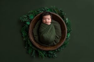 Newborn baby swaddled in green wrap sleeping in wooden bowl surrounded by lush green leaves on dark green background.