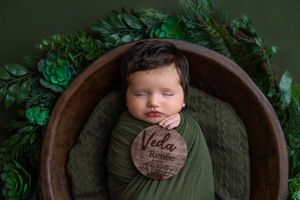 Newborn baby named Veda Renee sleeping peacefully in a wooden bowl wrapped in green, surrounded by lush foliage.