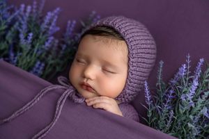 Sleeping newborn baby in purple knit bonnet wrapped in lavender blanket surrounded by purple lavender flowers.