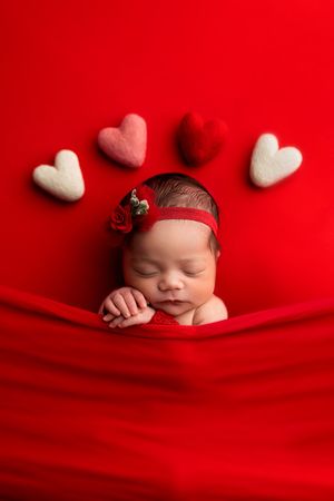 Sleeping newborn baby girl in red outfit with floral headband surrounded by felt hearts on red background.