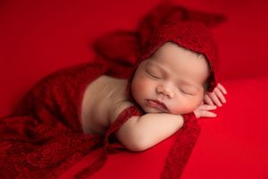 Newborn baby sleeping peacefully on red blanket wearing red lace bonnet in professional photography studio.