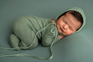 Sleeping newborn baby wrapped in sage green knit swaddle and bonnet on matching green background.