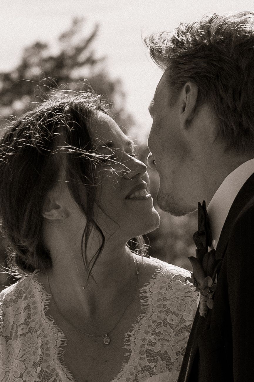 Black and white wedding photo of a smiling bride in lace dress and groom in suit sharing a romantic moment outdoors.