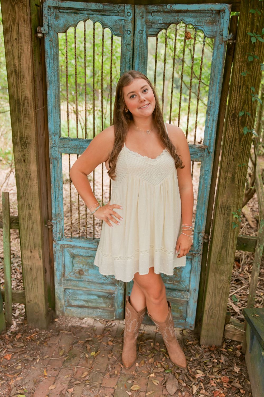 Young woman in white sundress and cowboy boots poses in front of rustic turquoise doors outdoors.