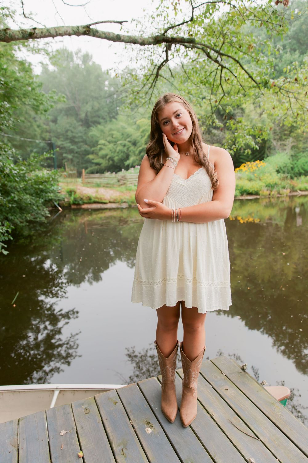 Young woman in white dress and cowboy boots smiling on wooden dock by serene pond surrounded by lush greenery.