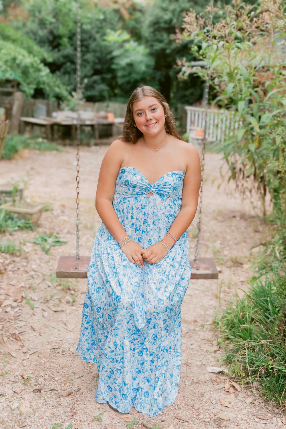 Young woman in blue floral strapless maxi dress smiling on a wooden swing in a garden setting.