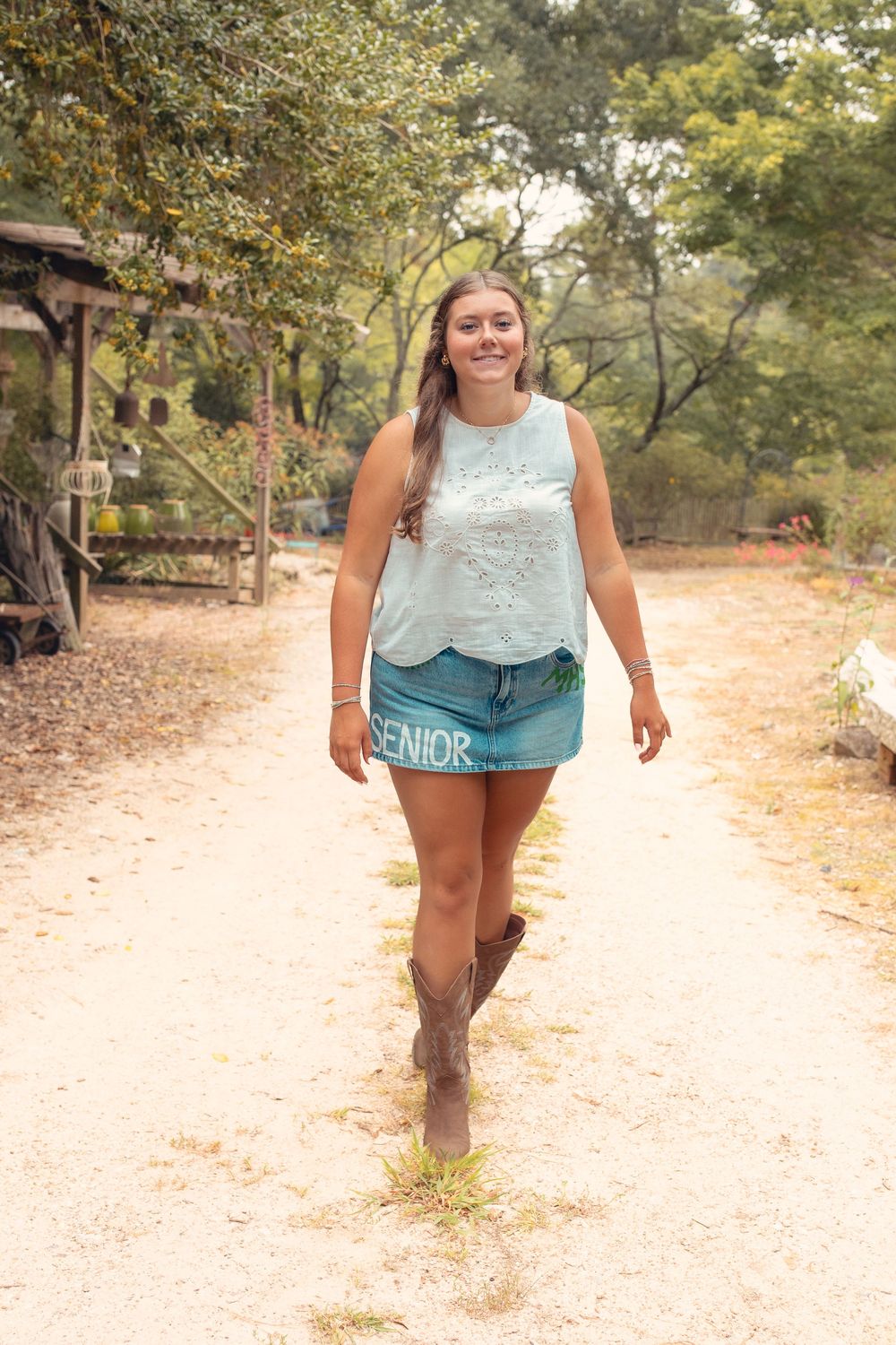 Smiling teenage girl in senior denim skirt, teal tank top, and cowboy boots walking on a sandy path surrounded by trees.