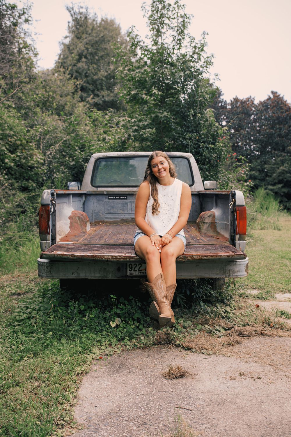 Young woman in white dress and boots sitting on rusty pickup truck tailgate surrounded by lush green trees.