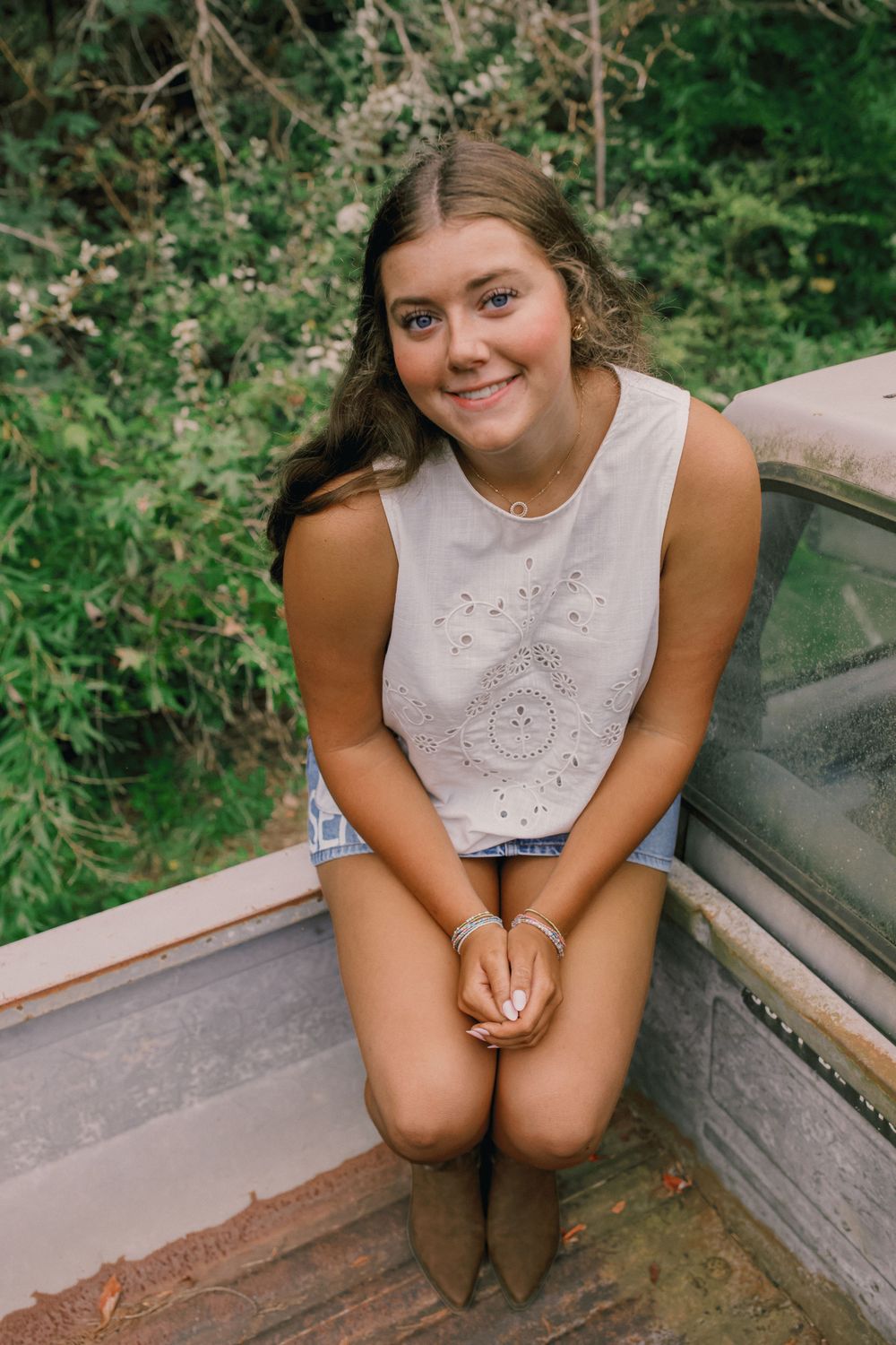 Young woman in white sleeveless top and denim shorts sits smiling on rustic wooden surface with green foliage background.