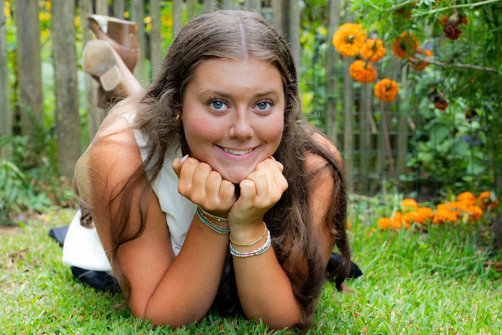 Smiling teenage girl lying on green grass in a garden with orange flowers and wooden fence in background.