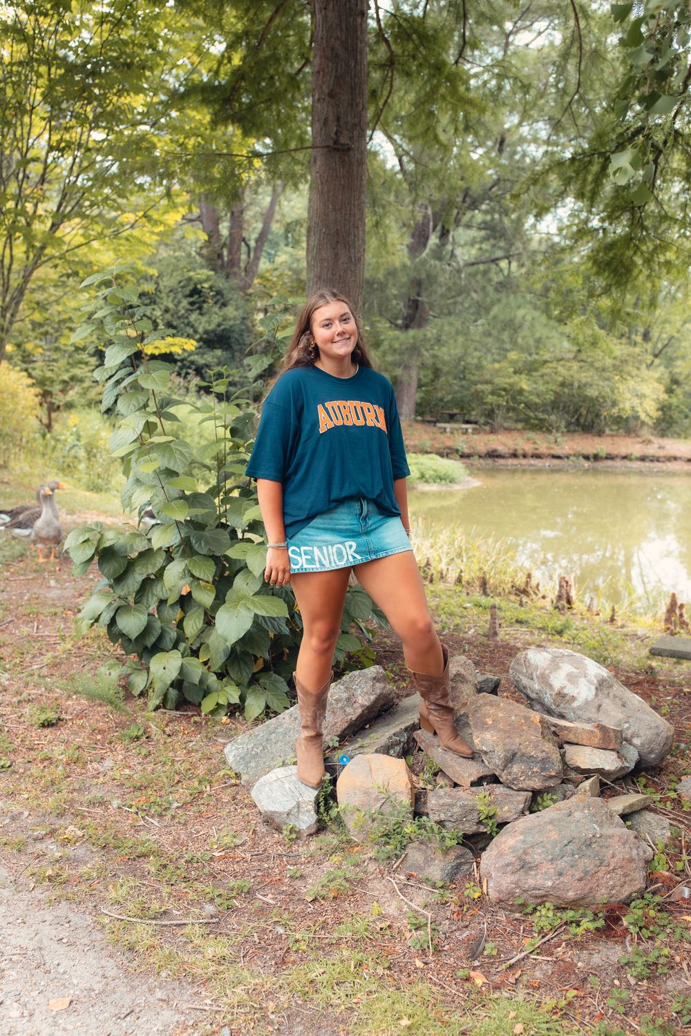 A smiling teenage girl in an Auburn teal shirt and shorts poses on rocks near a pond in a lush green park.