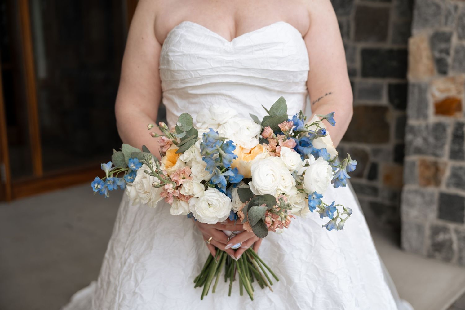 Bride in strapless white lace gown holding a romantic bouquet of white, peach, and blue flowers with eucalyptus.
