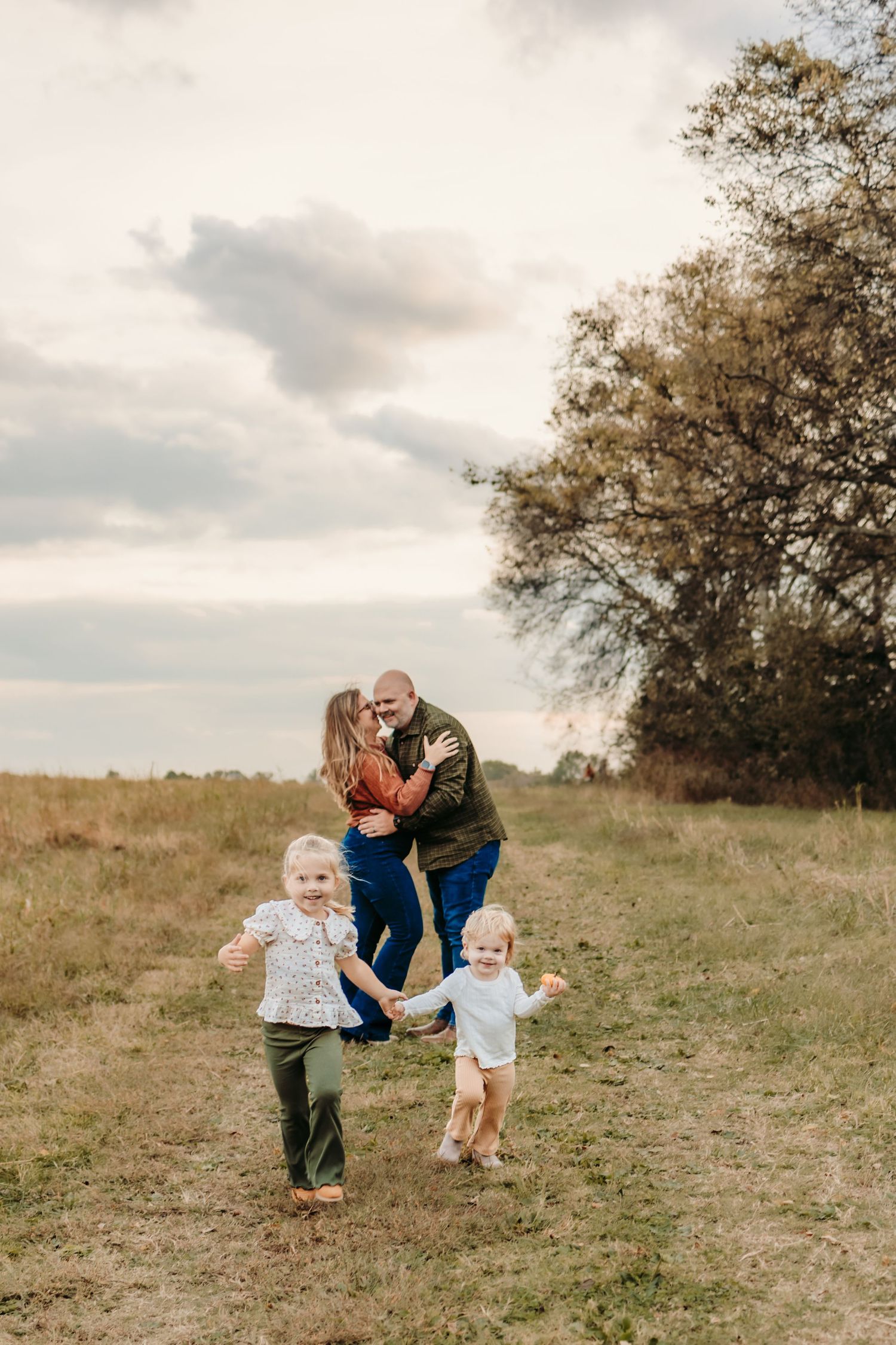 Family of four enjoying an outdoor autumn field, parents sharing a kiss while two young children play and hold hands.