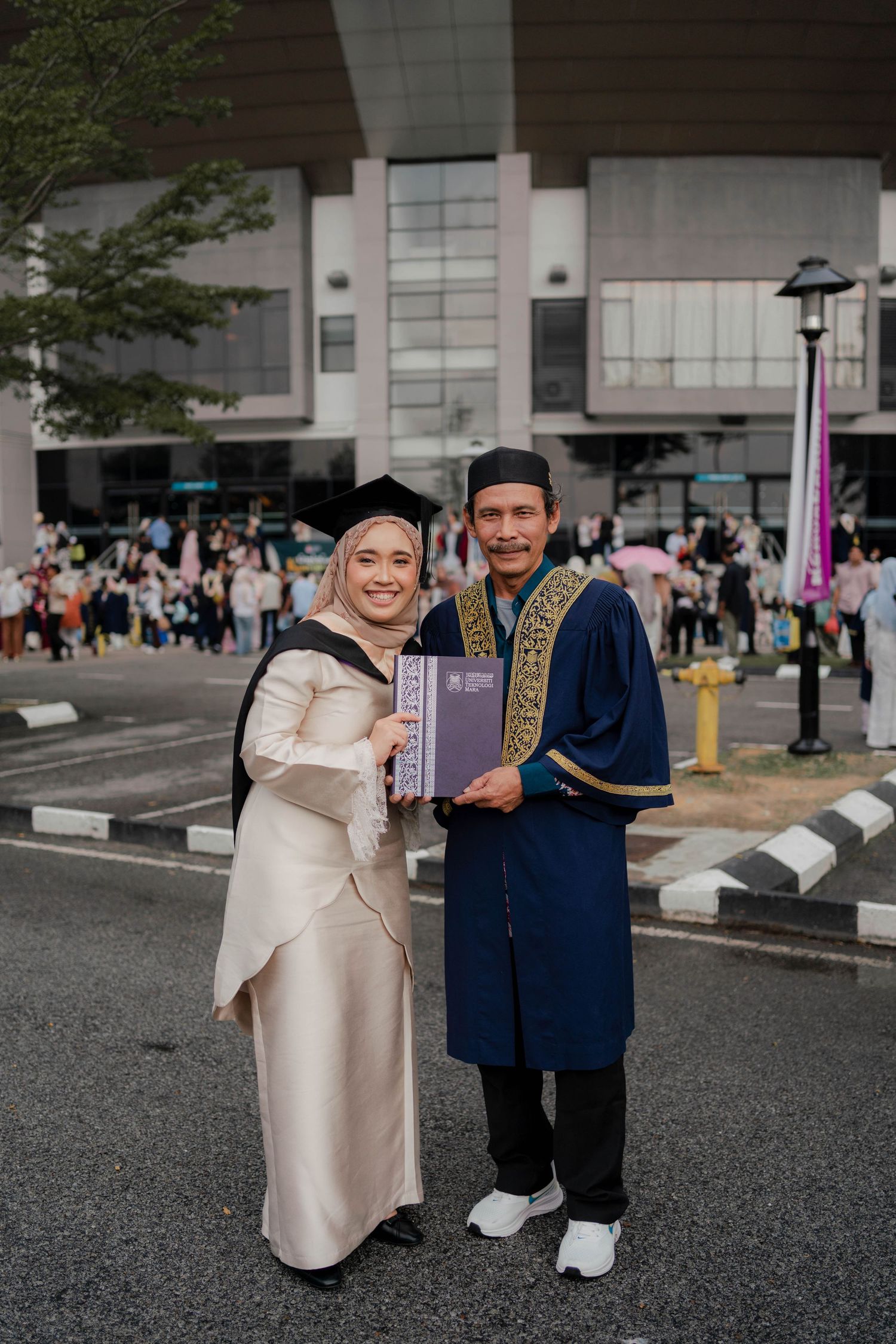 Muslim couple in traditional attire celebrating graduation, holding diploma outside university building.