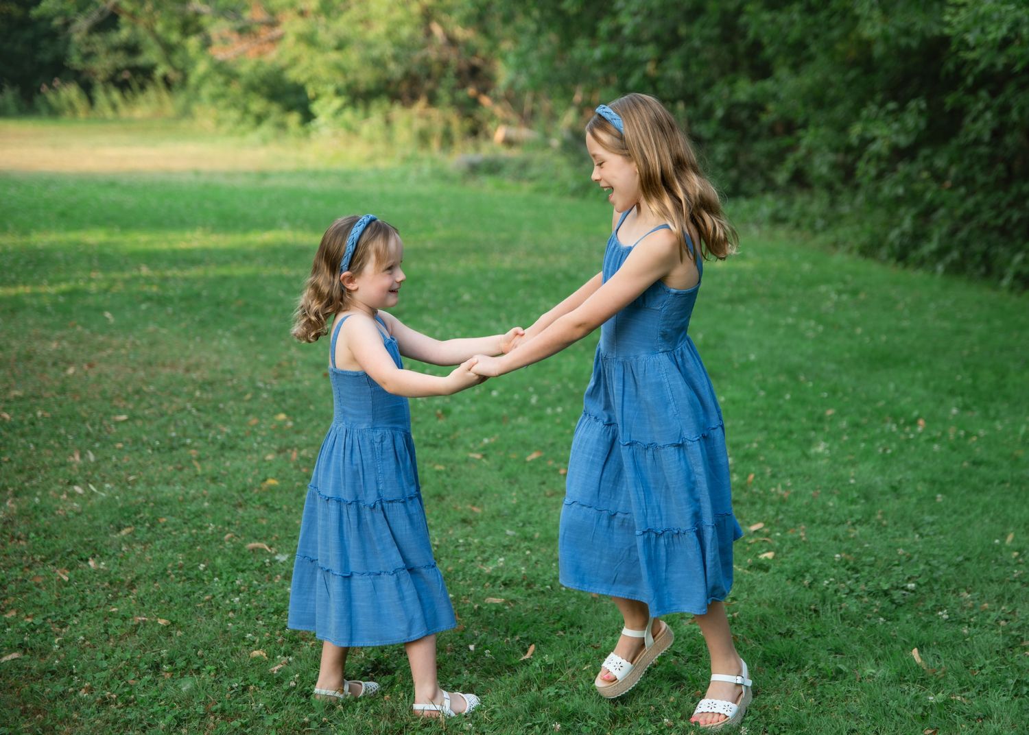 Two girls in matching blue dresses hold hands and dance together on a green lawn outdoors.