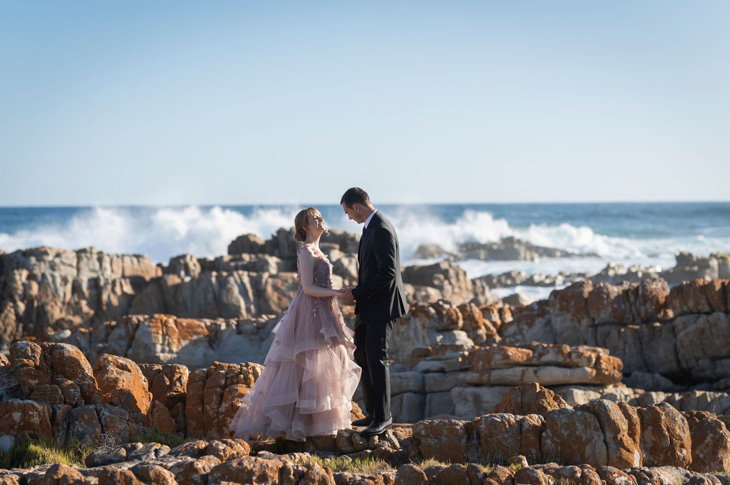 Couple in formal attire standing on rocky coastline with ocean waves crashing behind them on a clear day.