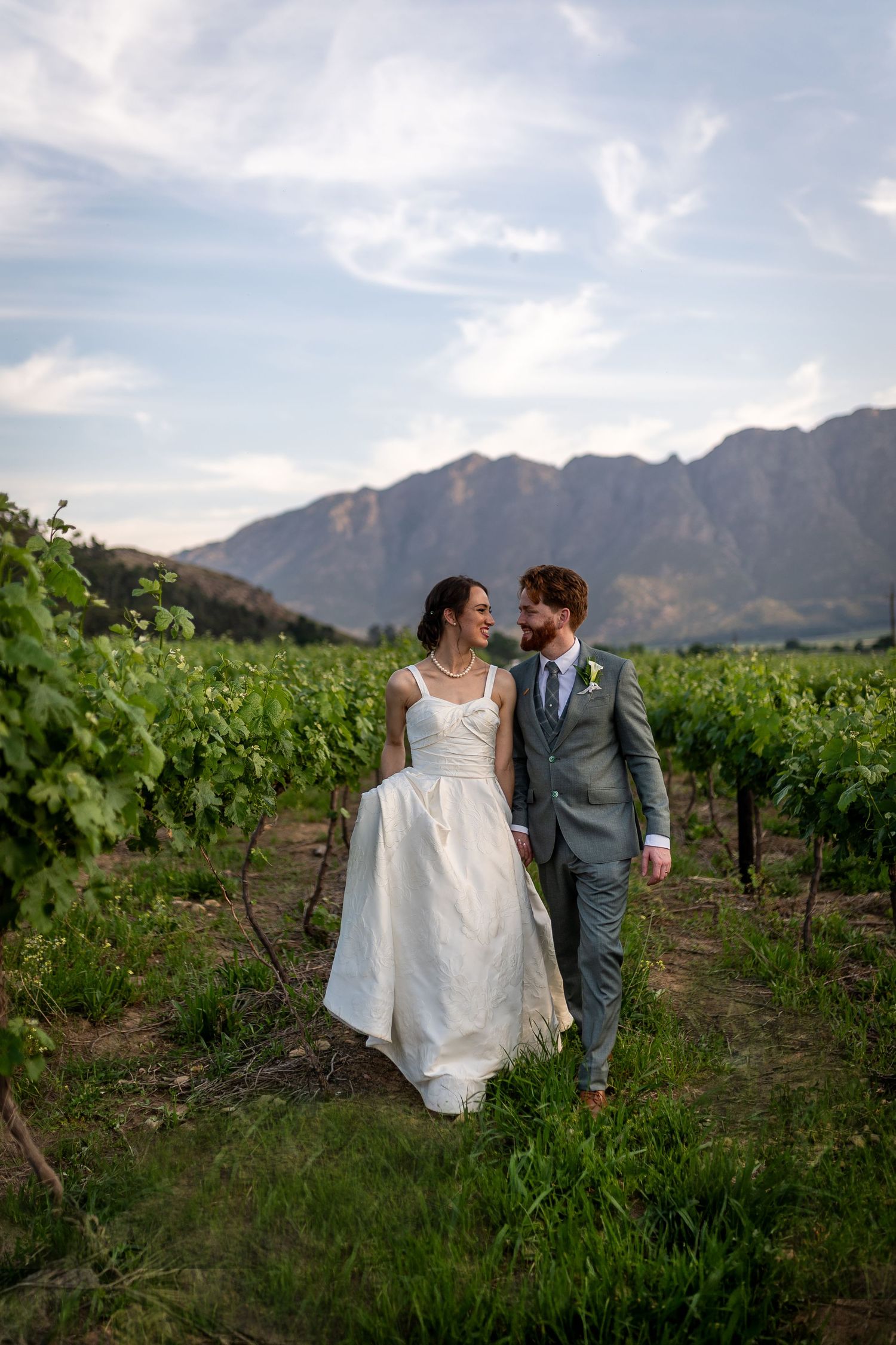 A bride and groom walk hand in hand through a lush vineyard with stunning mountain views on their wedding day.