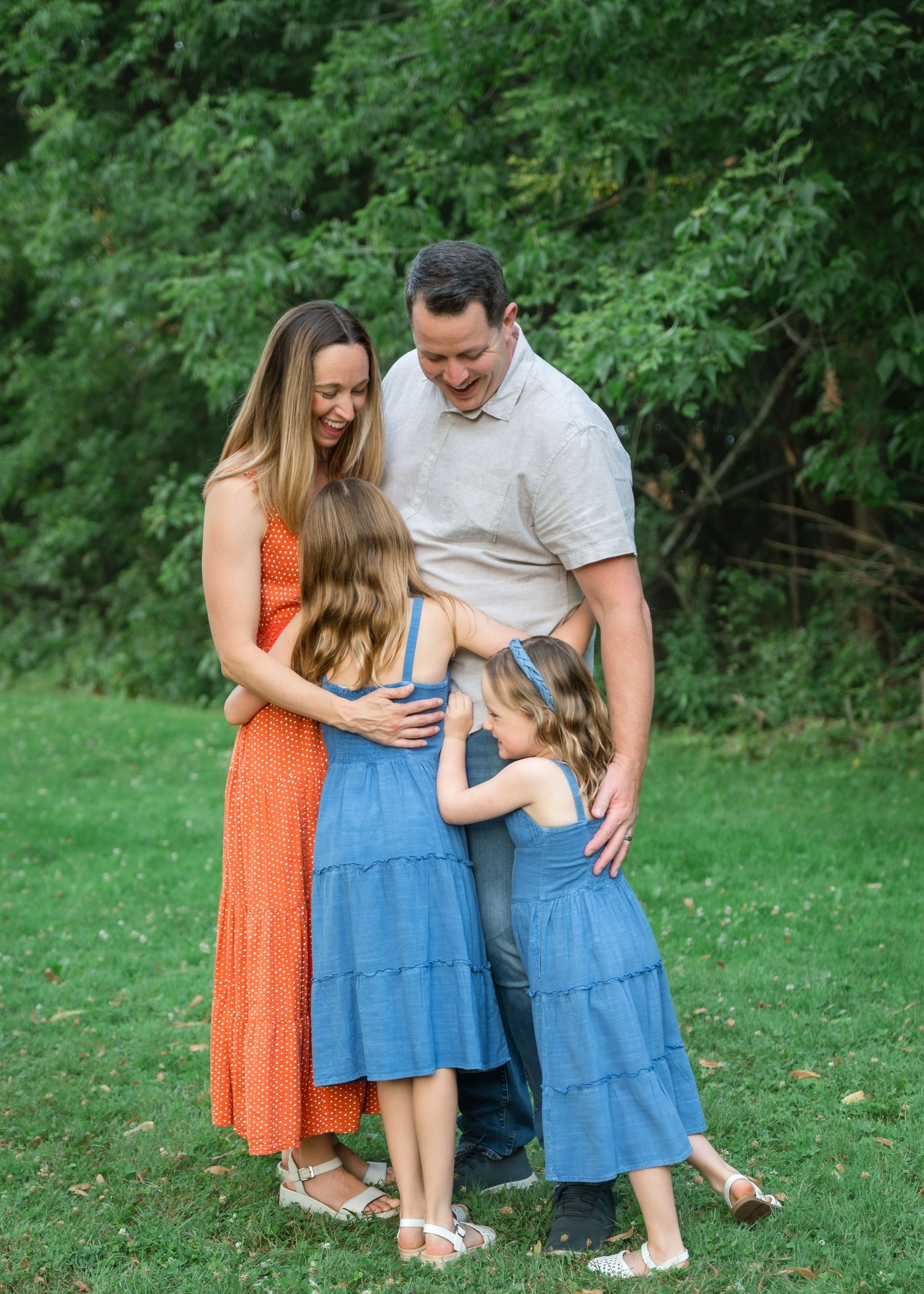 A joyful family of four shares a warm group hug on a green lawn with lush trees in the background.