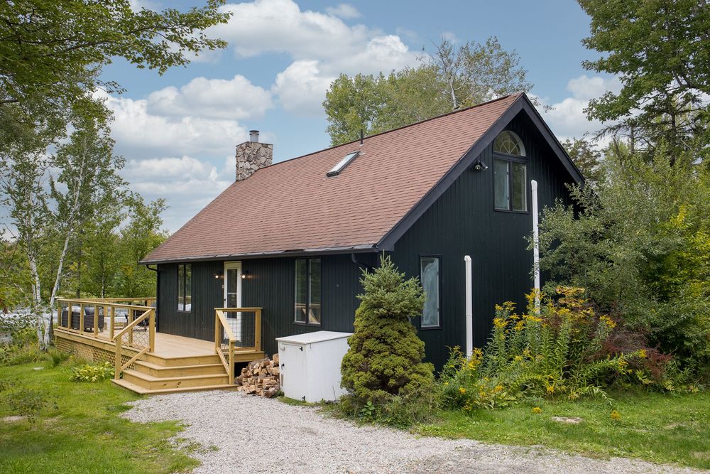 Dark green A-frame cottage with wood deck, stone chimney, skylights, and landscaped gravel driveway.