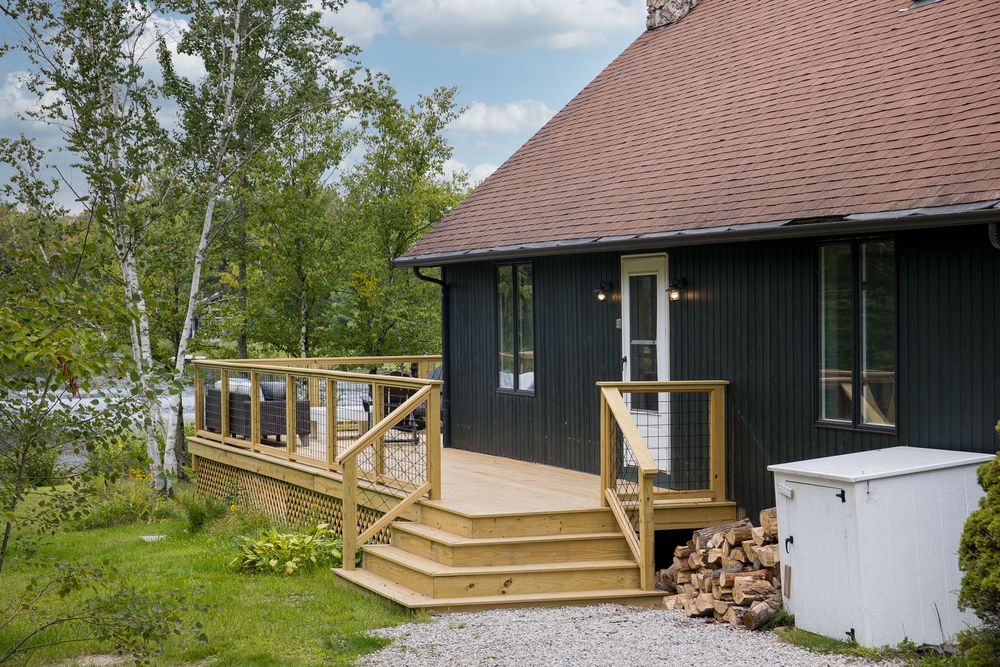 Dark green cottage exterior with new wooden deck, cable railing, stacked firewood, and birch trees.