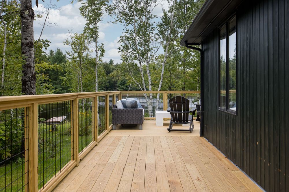 New wooden deck with cable railing, Adirondack chairs, outdoor seating, and birch trees overlooking yard.