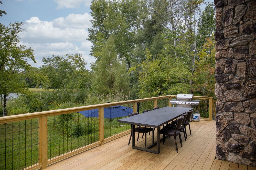 Outdoor dining table on wooden deck with stone pillar and gas grill beside lush greenery.
