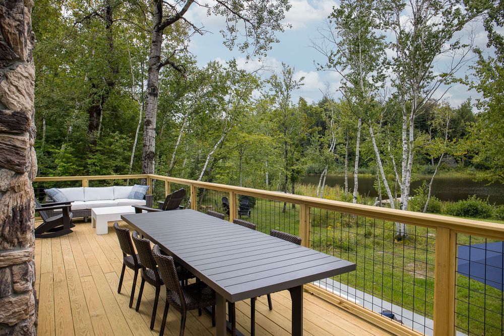 Dark dining table set on wood deck with stone chimney and lake views through birch trees.