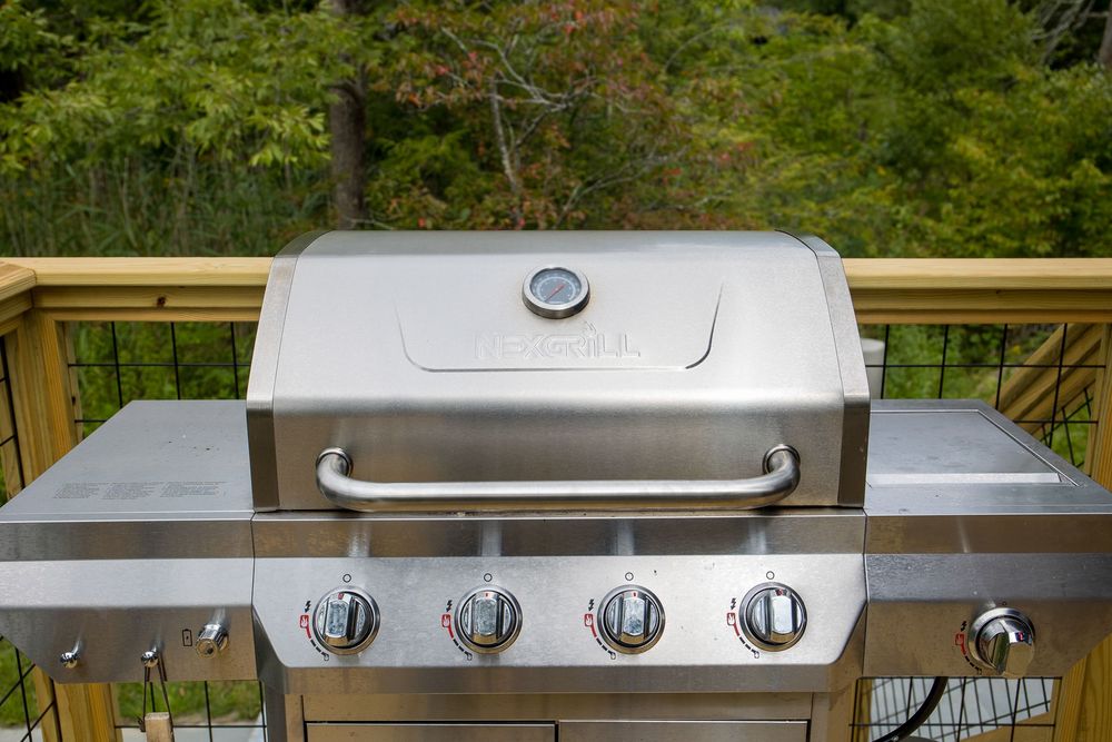 Stainless steel four-burner gas grill on outdoor deck with lush green trees in background.