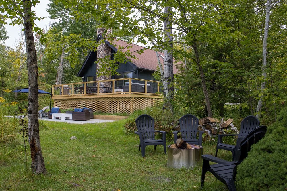 Black Adirondack chairs around fire pit with wooden deck and cottage visible through green trees.