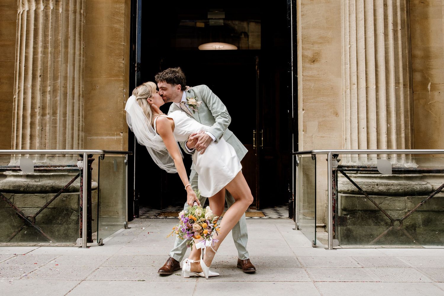 A smiling couple in wedding attire poses in a charming stone alleyway, groom in light gray suit, bride in white mini dress.