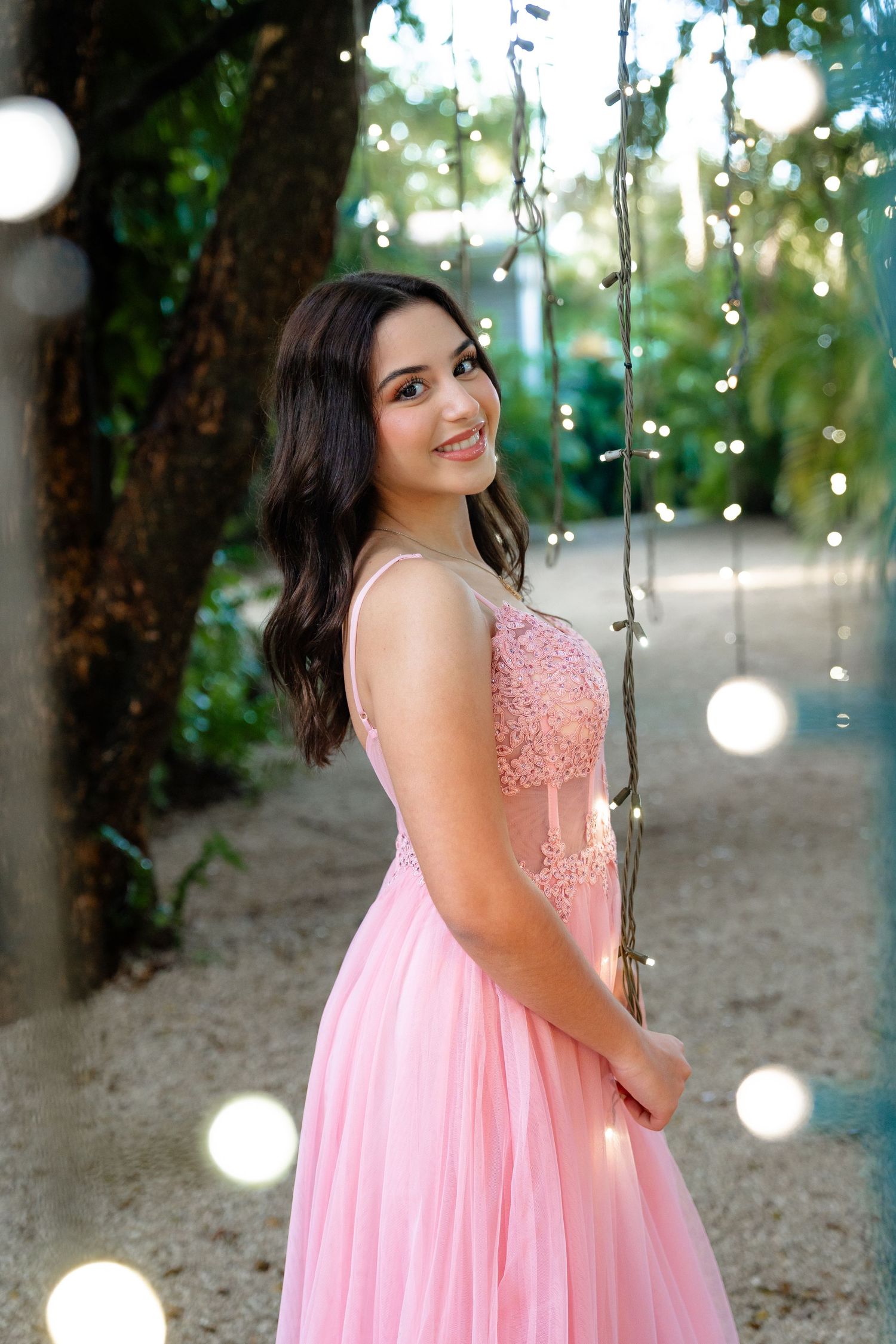 Young woman in pink lace ball gown smiling outdoors among twinkling string lights and lush greenery.