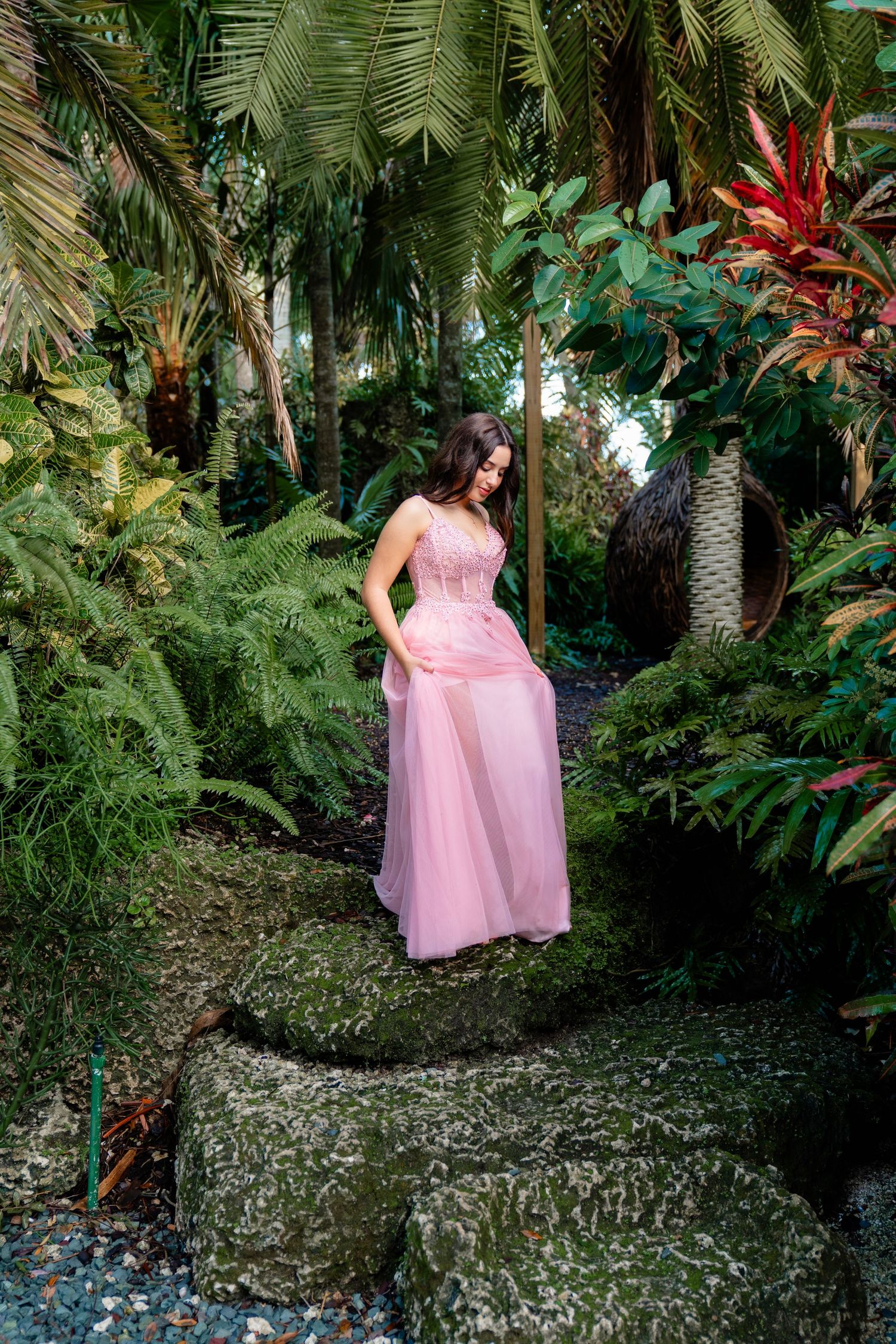 Woman in pink gown standing on mossy steps surrounded by lush tropical garden with ferns and palm trees.