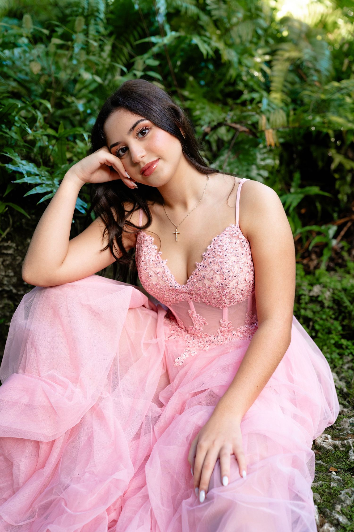 Young woman in pink beaded tulle ball gown poses gracefully outdoors among lush green ferns.