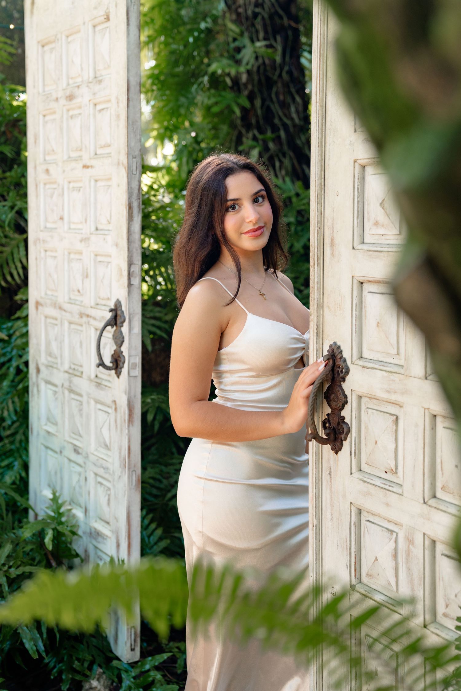 Young woman in elegant satin slip dress standing between rustic white doors surrounded by lush green foliage.