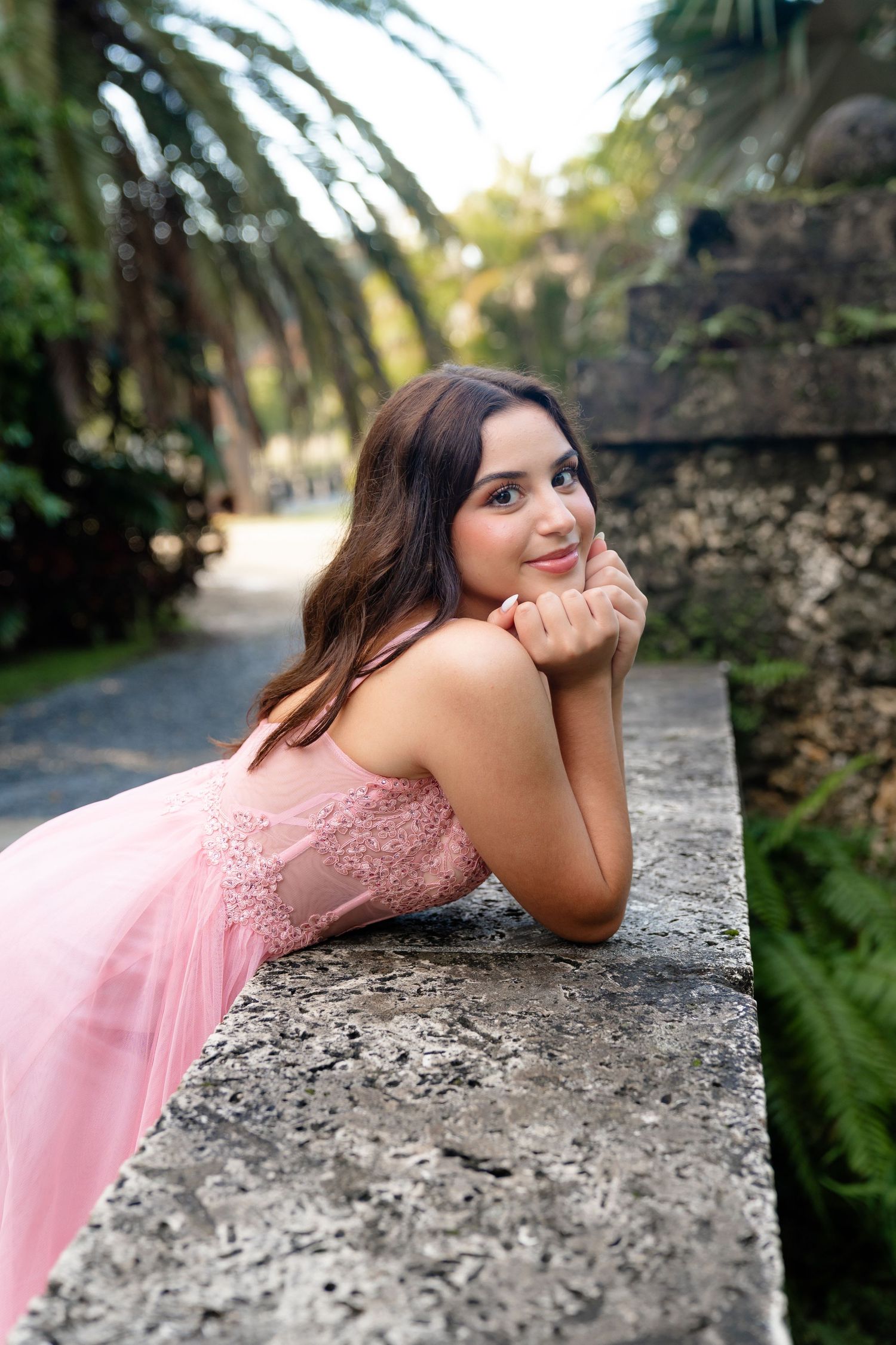 Young woman in pink dress leaning on stone wall, smiling in lush garden with palm trees.