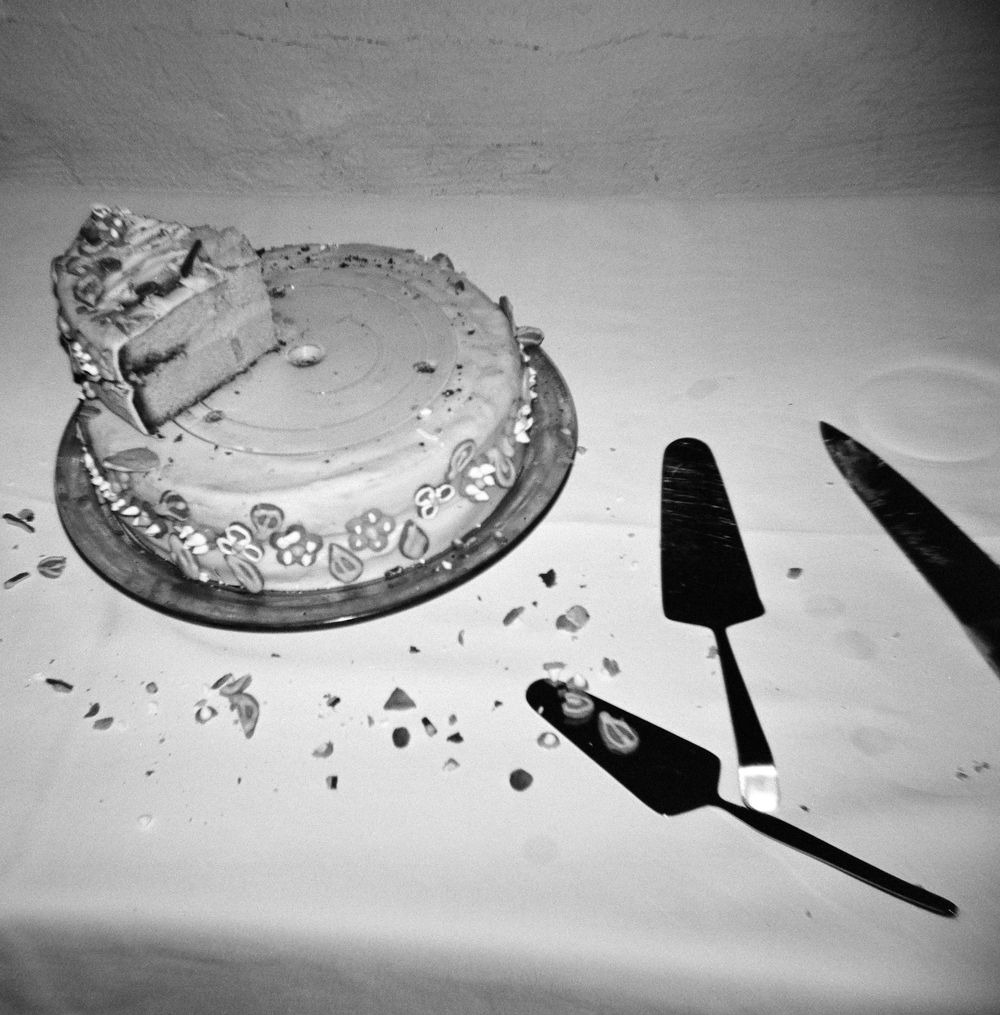 Black and white photo of a partially sliced frosted cake on a stand with a cake server and knife on a white tablecloth.