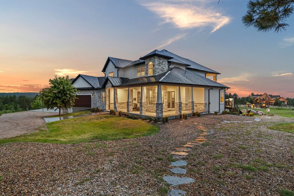 Stunning two-story home with wraparound porch and stone accents photographed at sunset with gravel driveway.