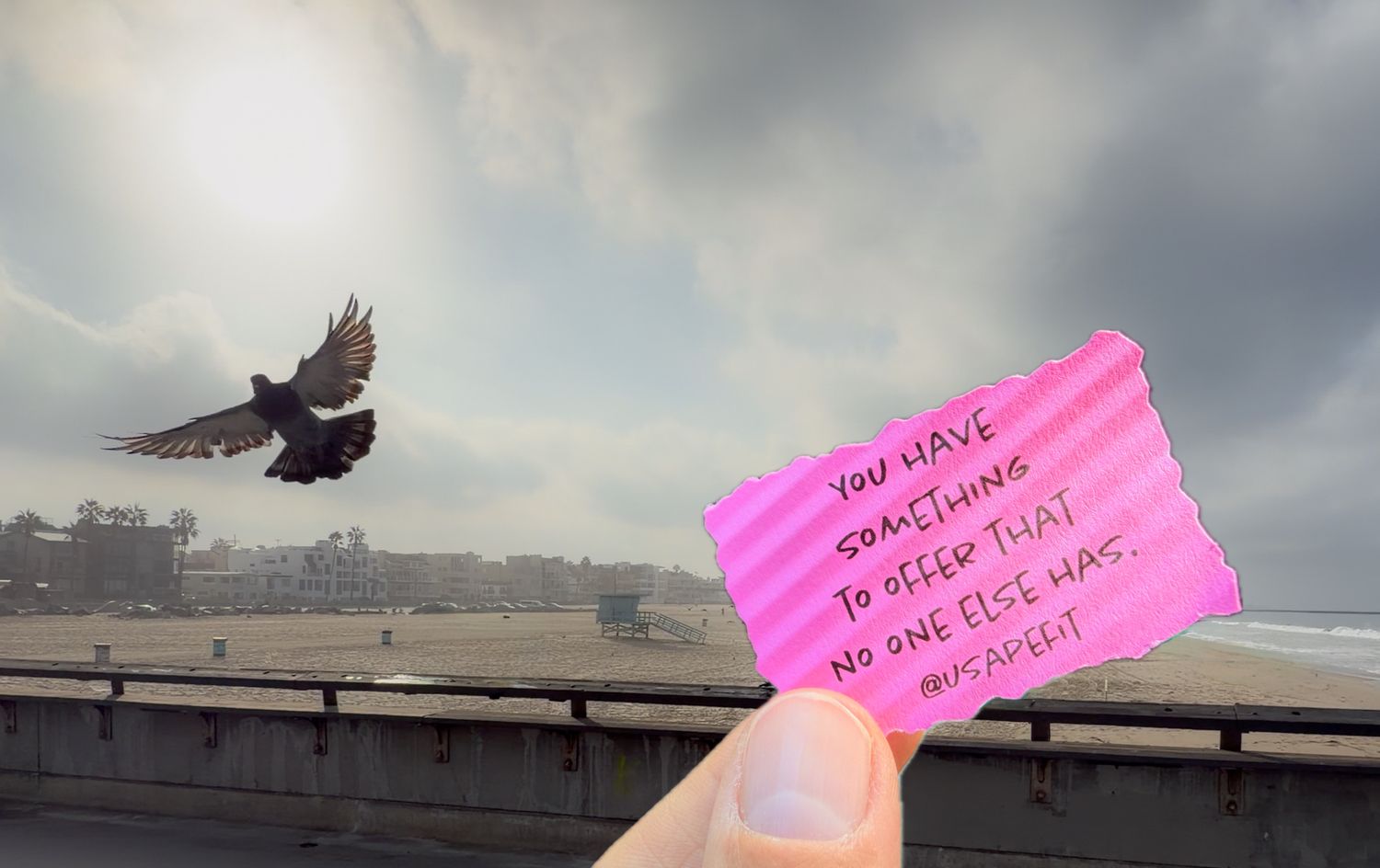 Hand holding pink motivational note near ocean with bird in flight against cloudy sky backdrop.